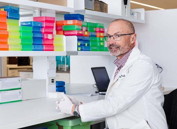 Professor David Mackey wearing a white lab coat and white disposable gloves, sitting at a bench in the research lab and facing the camera, with colourful boxes on a shelf nearby.